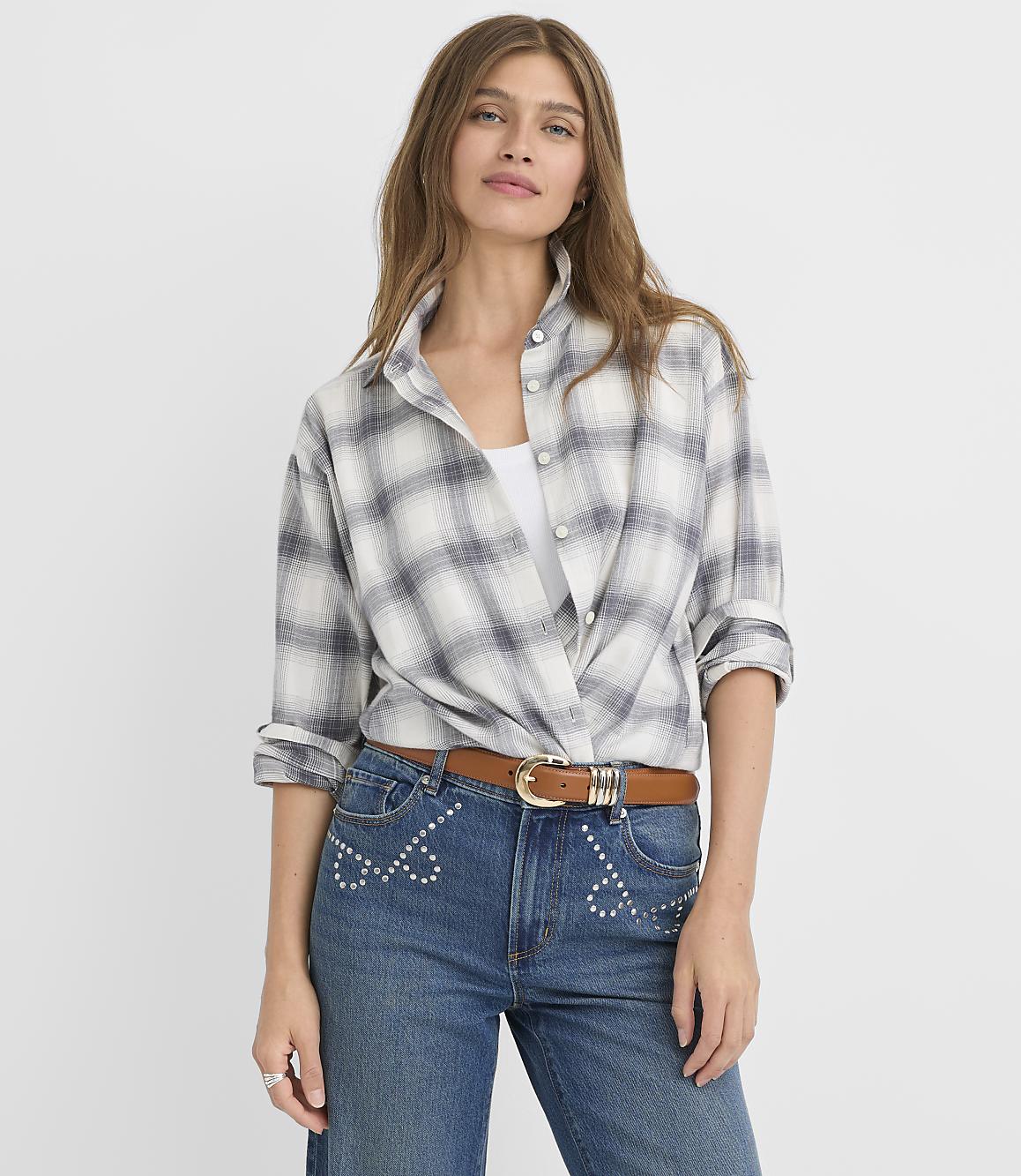 A woman in a plaid shirt and jeans stands against a plain white background.