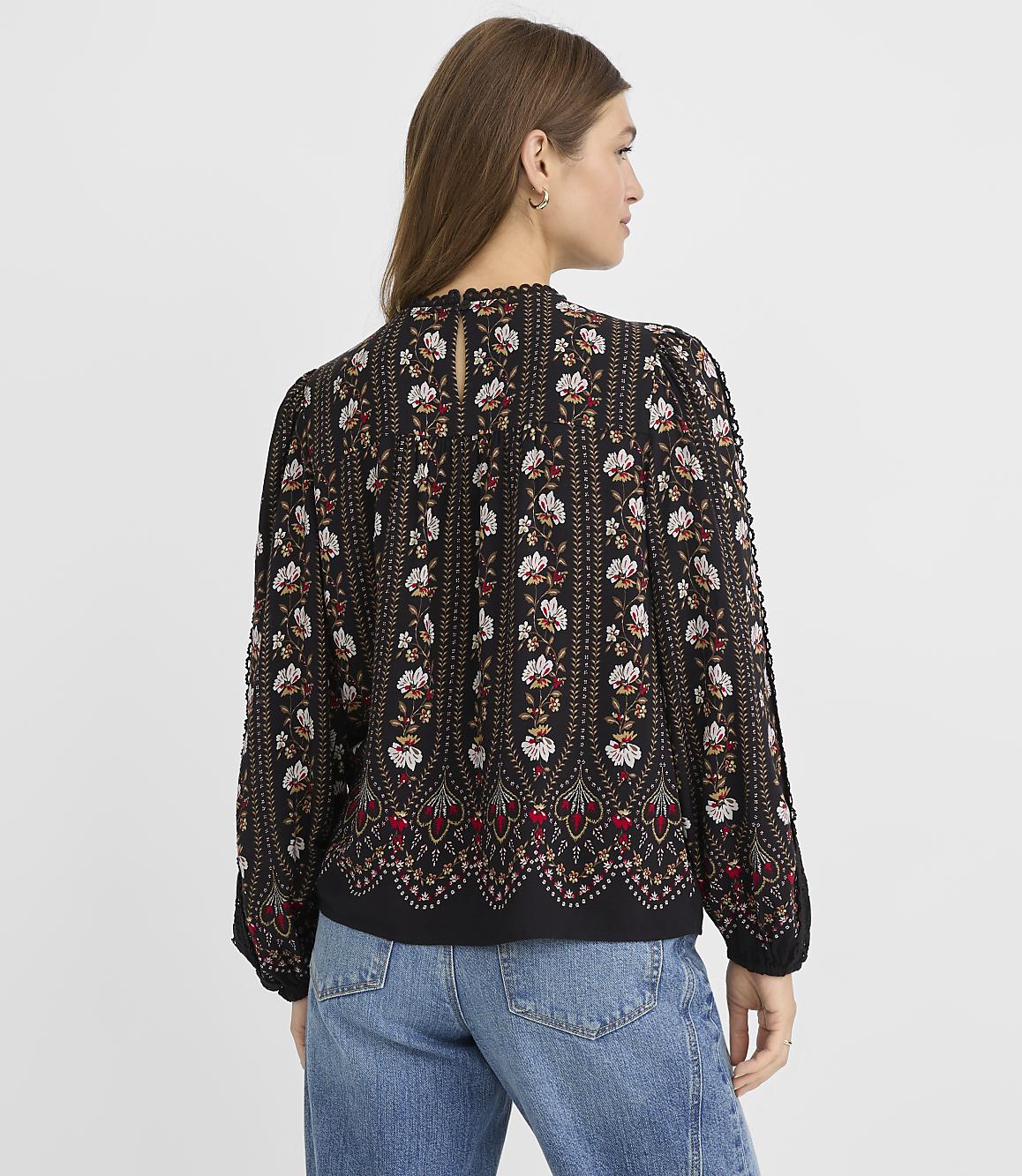 A woman in a patterned blouse and jeans stands against a plain gray background.