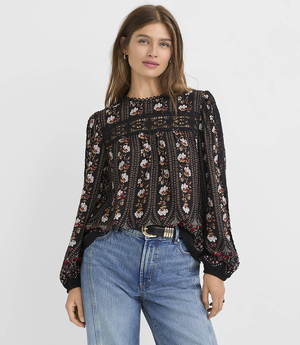 A woman in a floral blouse and jeans stands against a plain gray background.