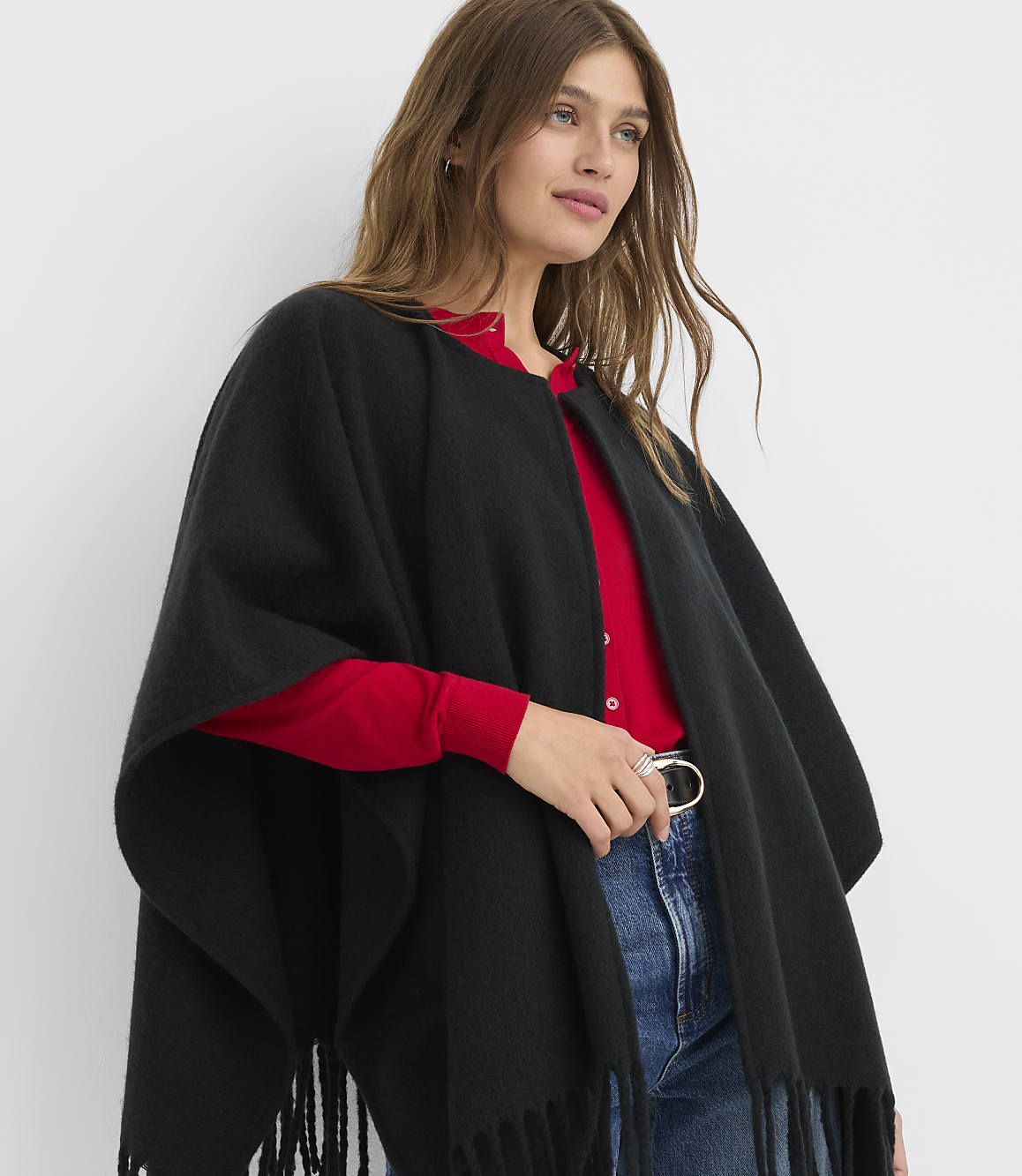 A woman in a black fringed poncho over a red top and blue jeans stands against a plain white background.