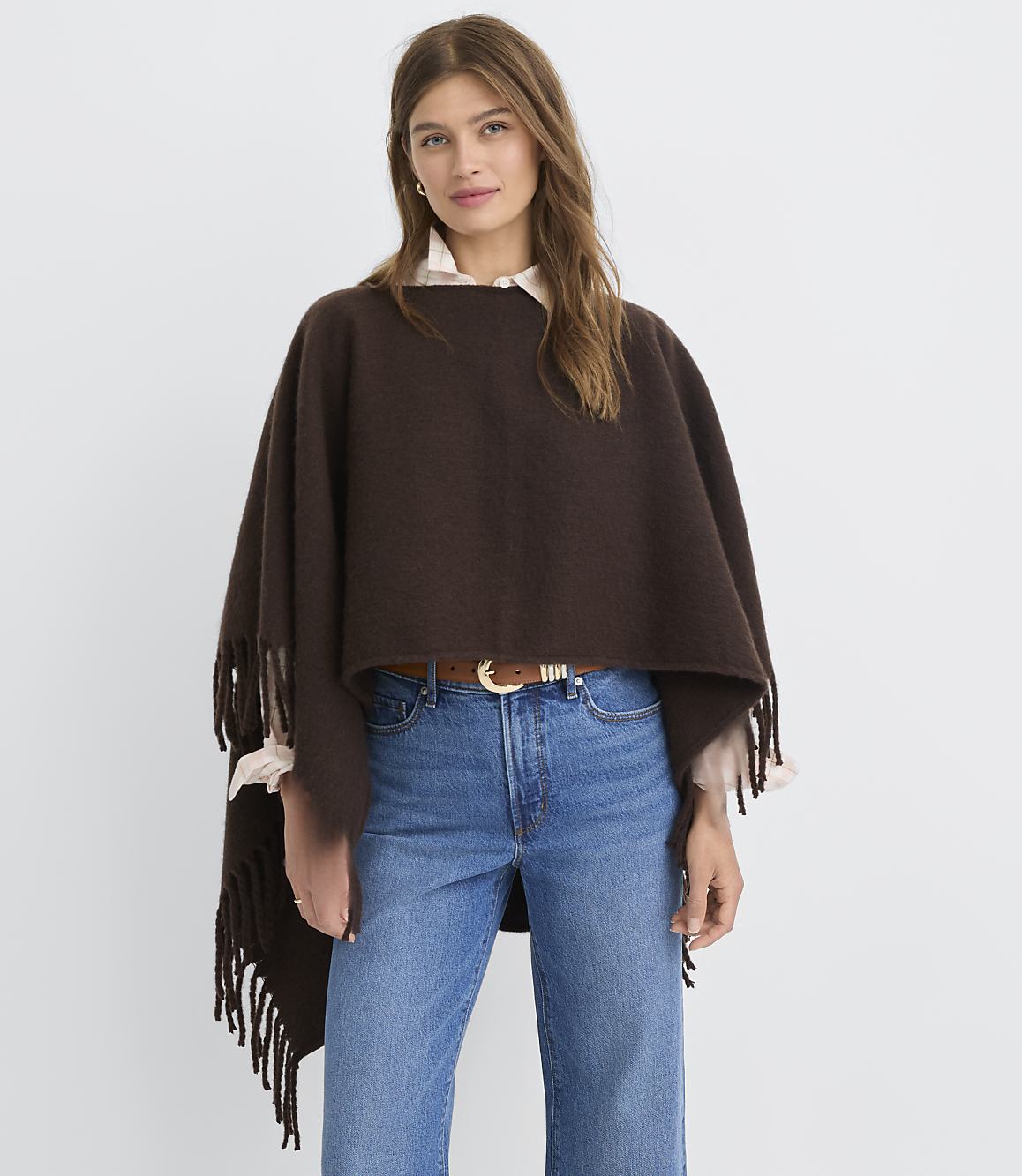 A woman in a brown fringed poncho and blue jeans stands against a plain white background.