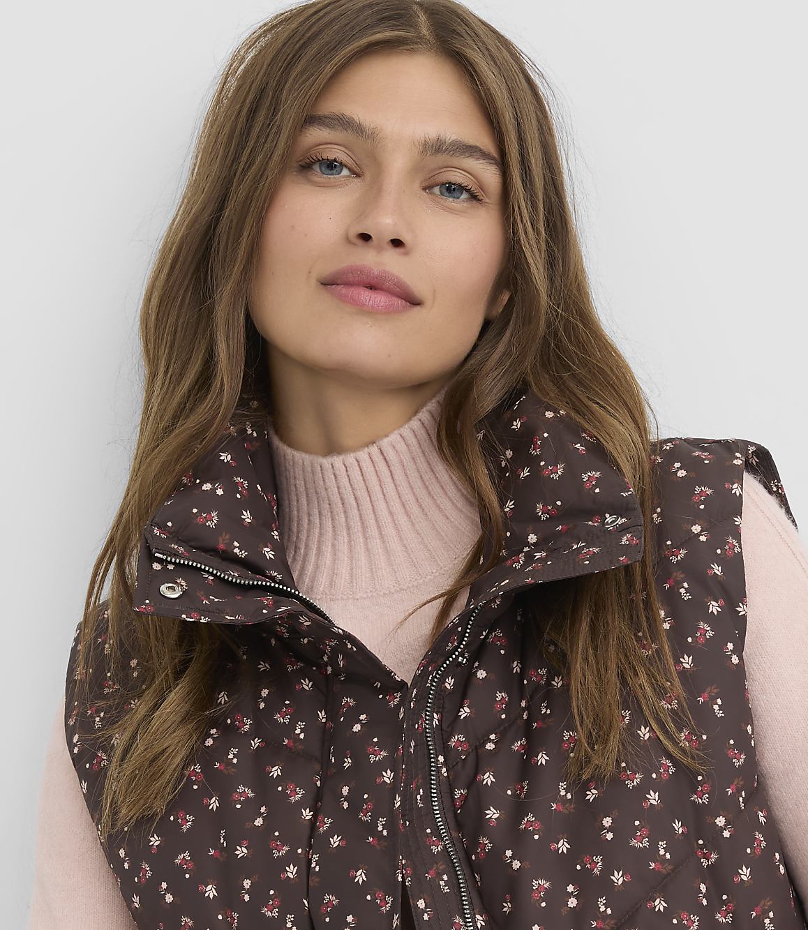 A woman in a floral vest and pink turtleneck looks forward against a plain background.