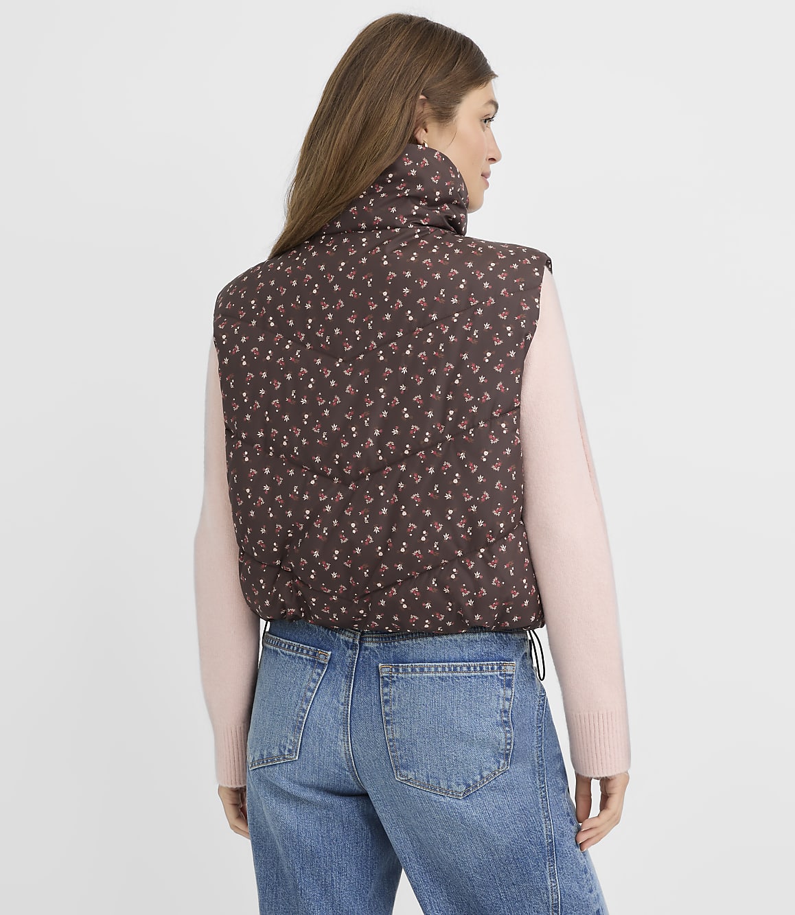 A woman in a floral puffer vest and pink sweater stands against a plain white background.