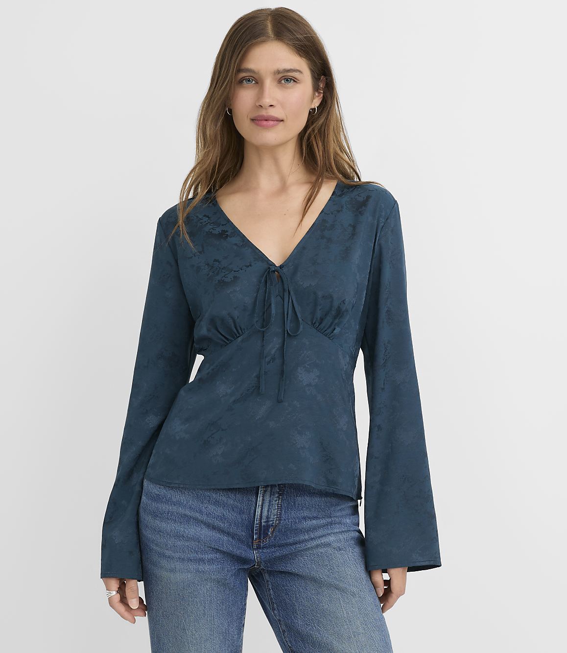 A woman in a teal, long-sleeved blouse and blue jeans stands against a plain white background.