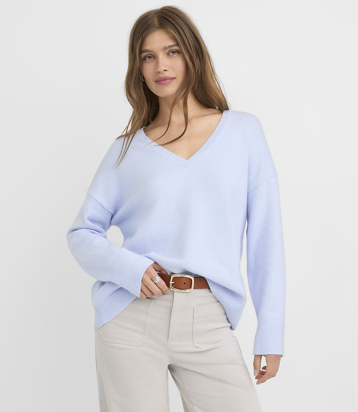 A woman in a light blue v-neck sweater and tan pants stands against a plain white background.