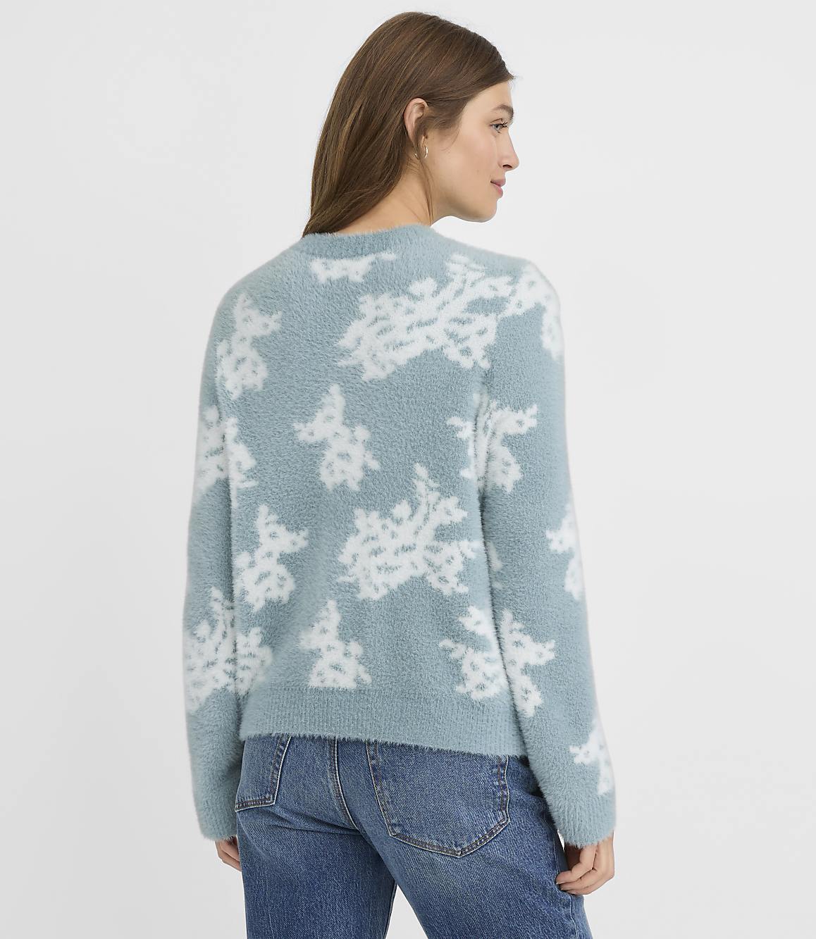 A woman in a fuzzy blue sweater with a white floral pattern stands against a plain, light blue background.