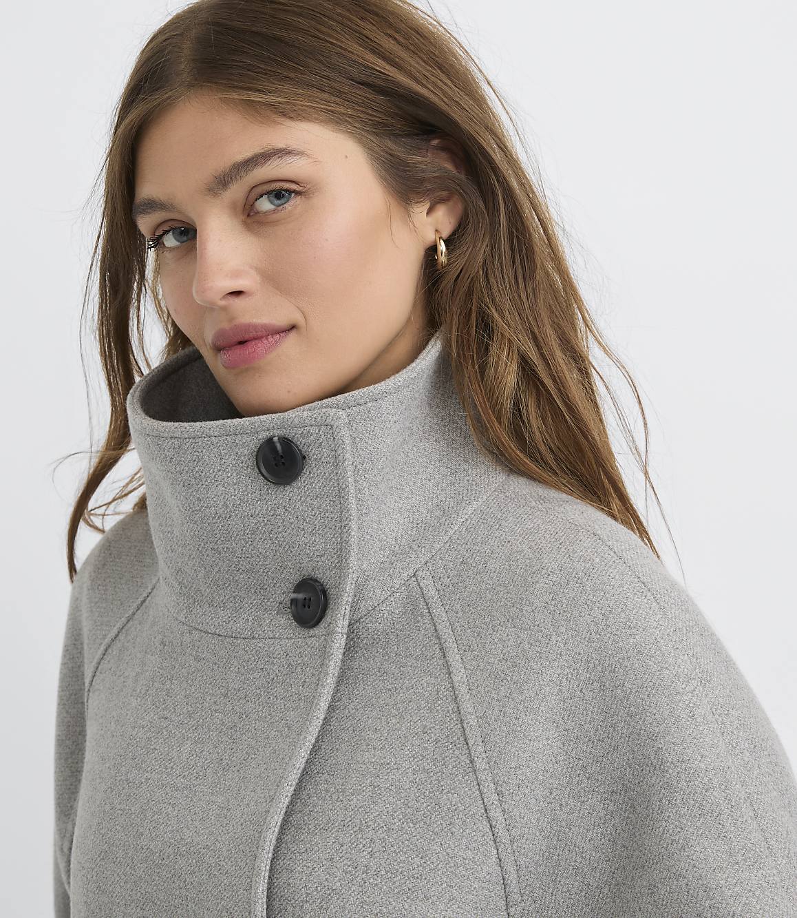 A woman with light brown hair and blue eyes, wearing a gray coat with a high collar and two black buttons, is shown against a plain gray background.