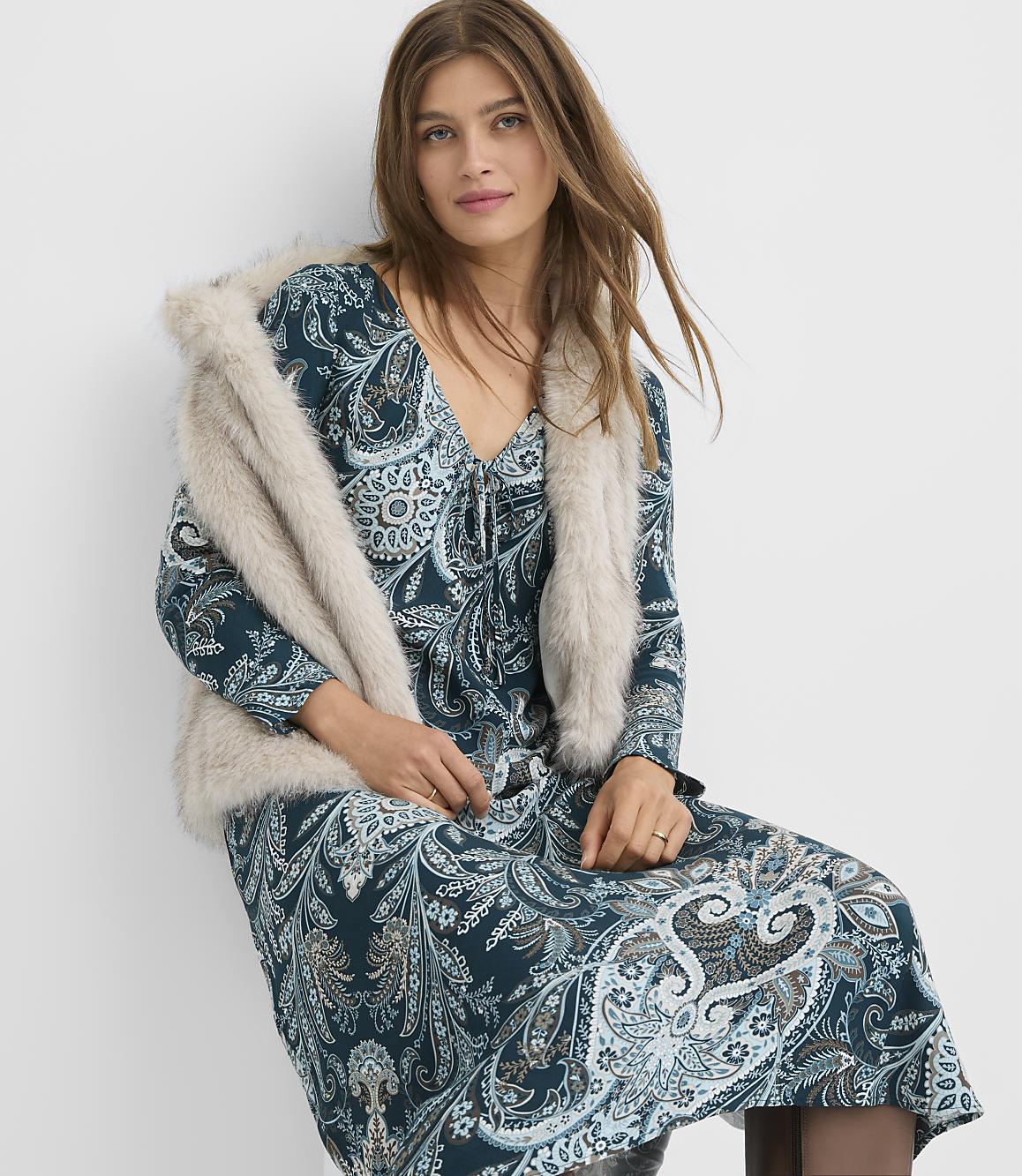 A woman in a teal paisley dress and a faux fur stole poses against a plain white background.