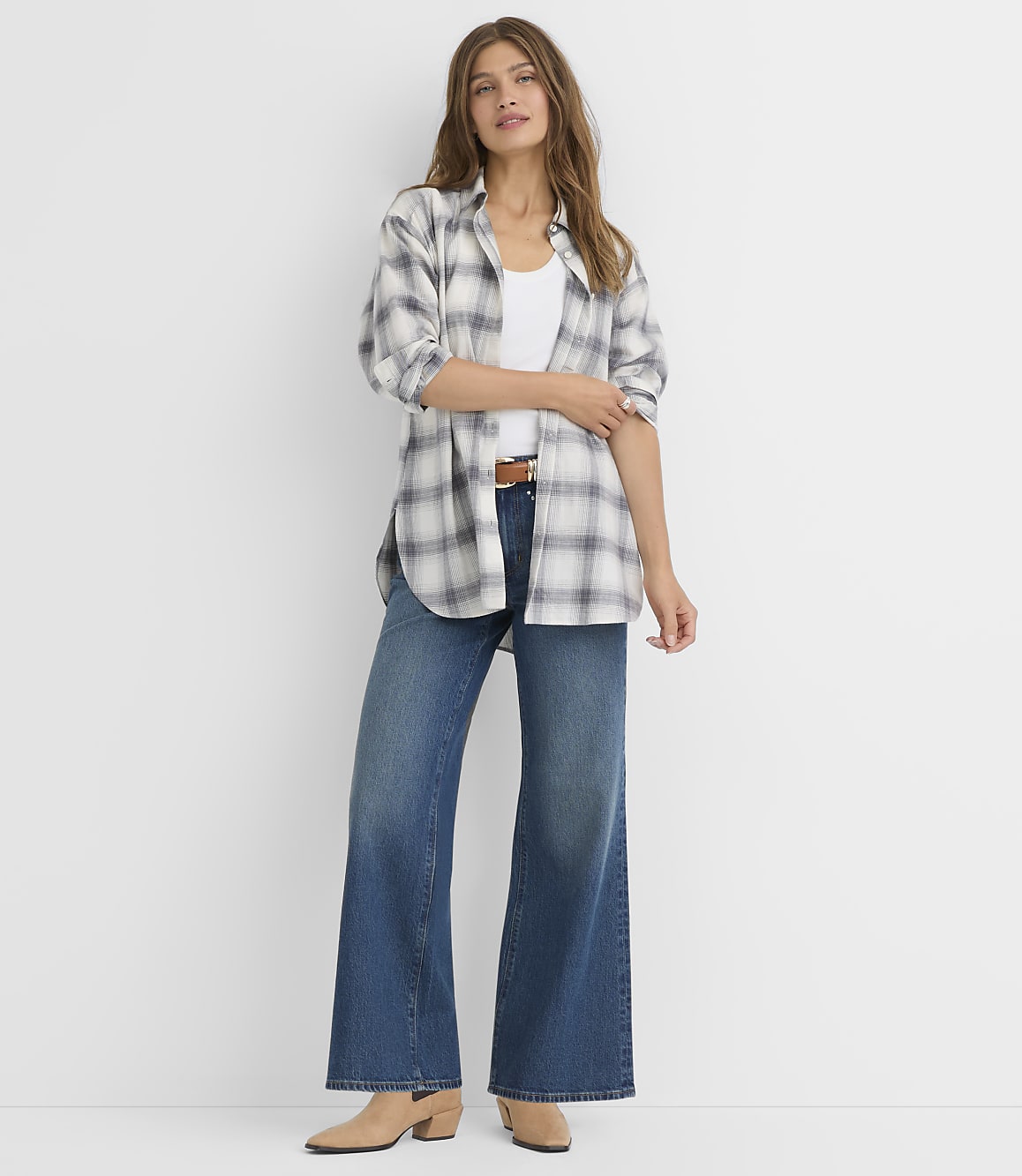 A woman in a plaid shirt and wide-leg jeans stands against a plain white background.