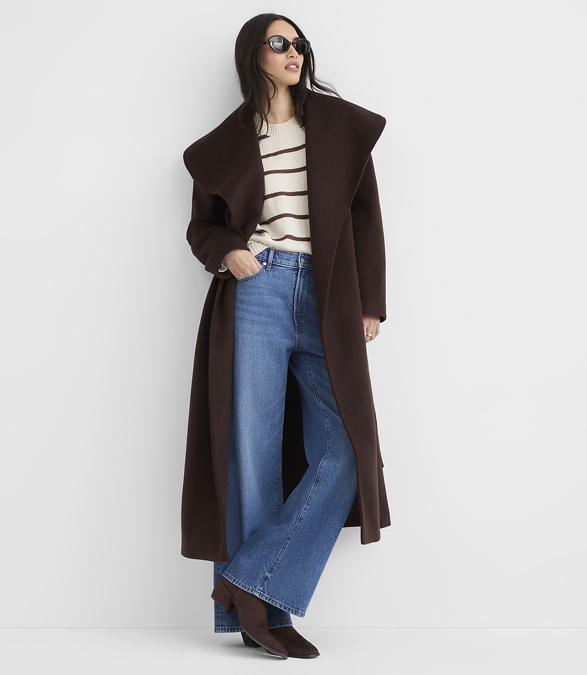 A woman in a long brown coat and wide-leg jeans stands against a plain white background.