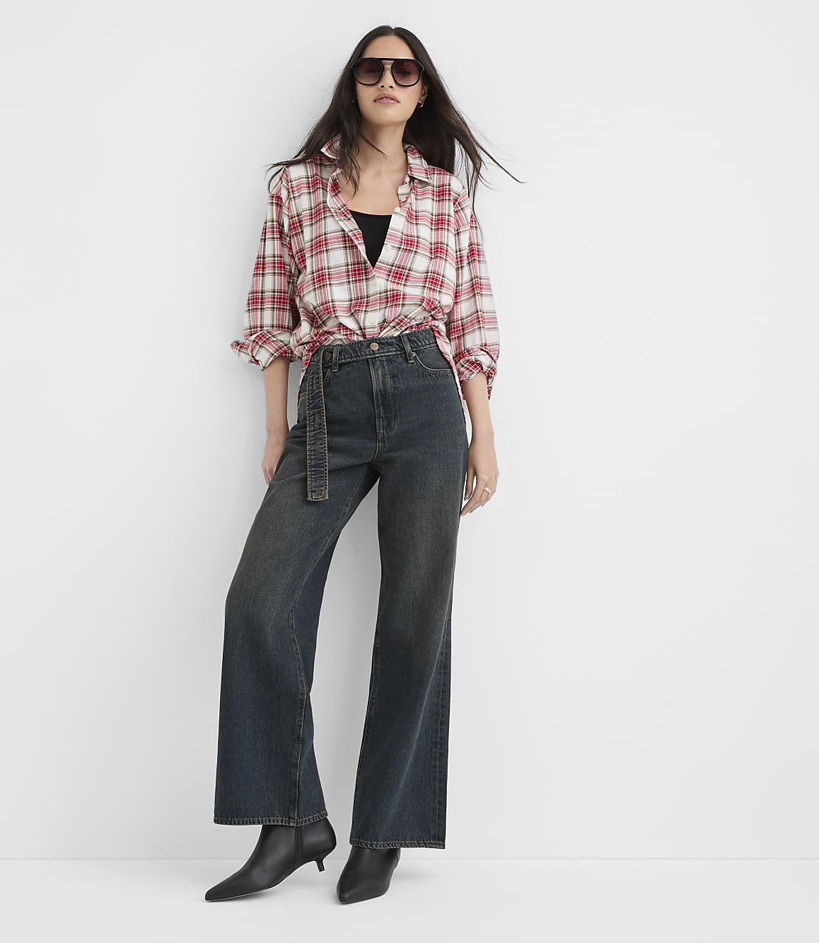A woman in a red and white plaid shirt and dark wide-leg jeans stands against a plain white background.