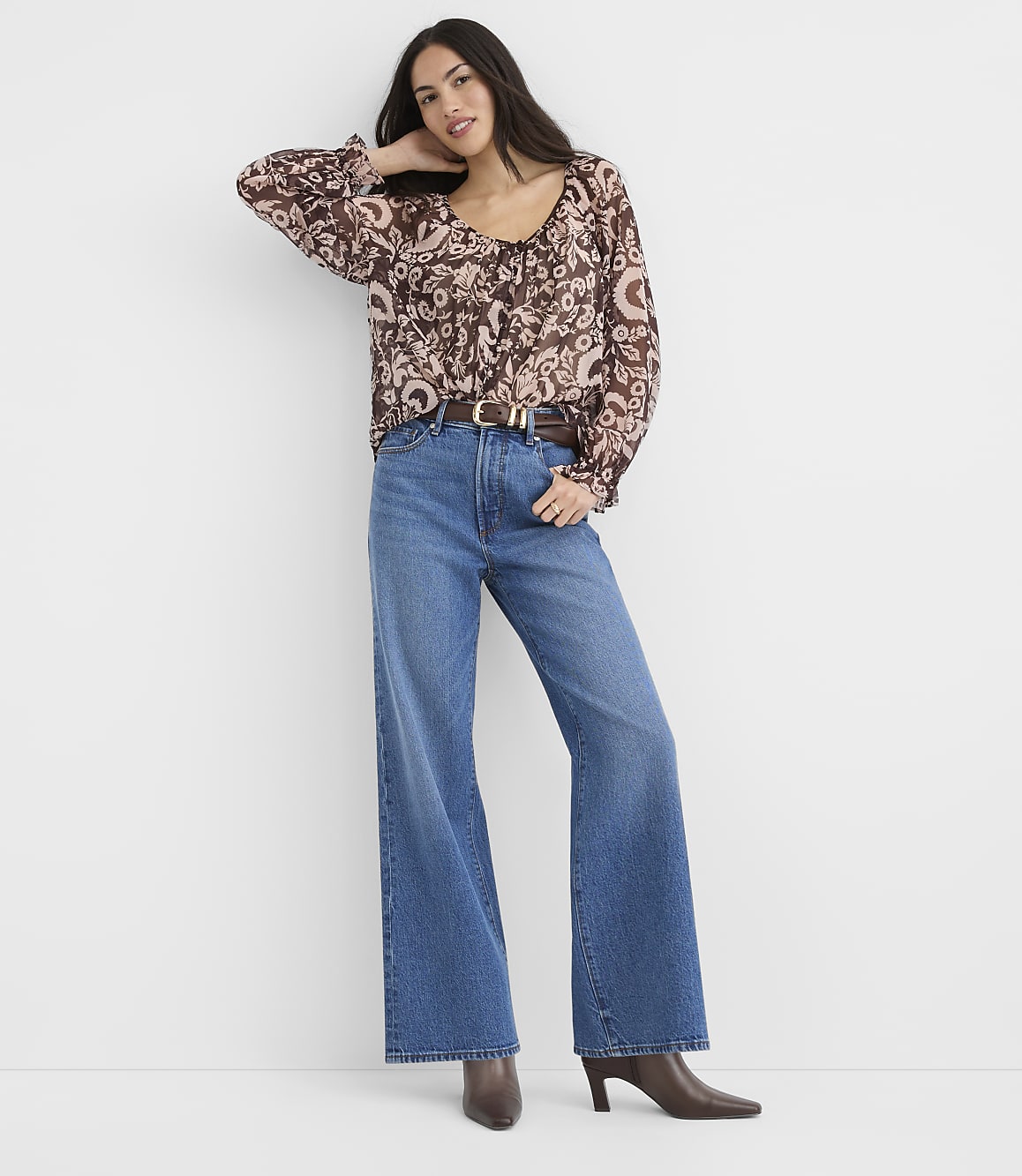 A woman in a patterned brown blouse and wide-leg blue jeans stands against a plain white background.