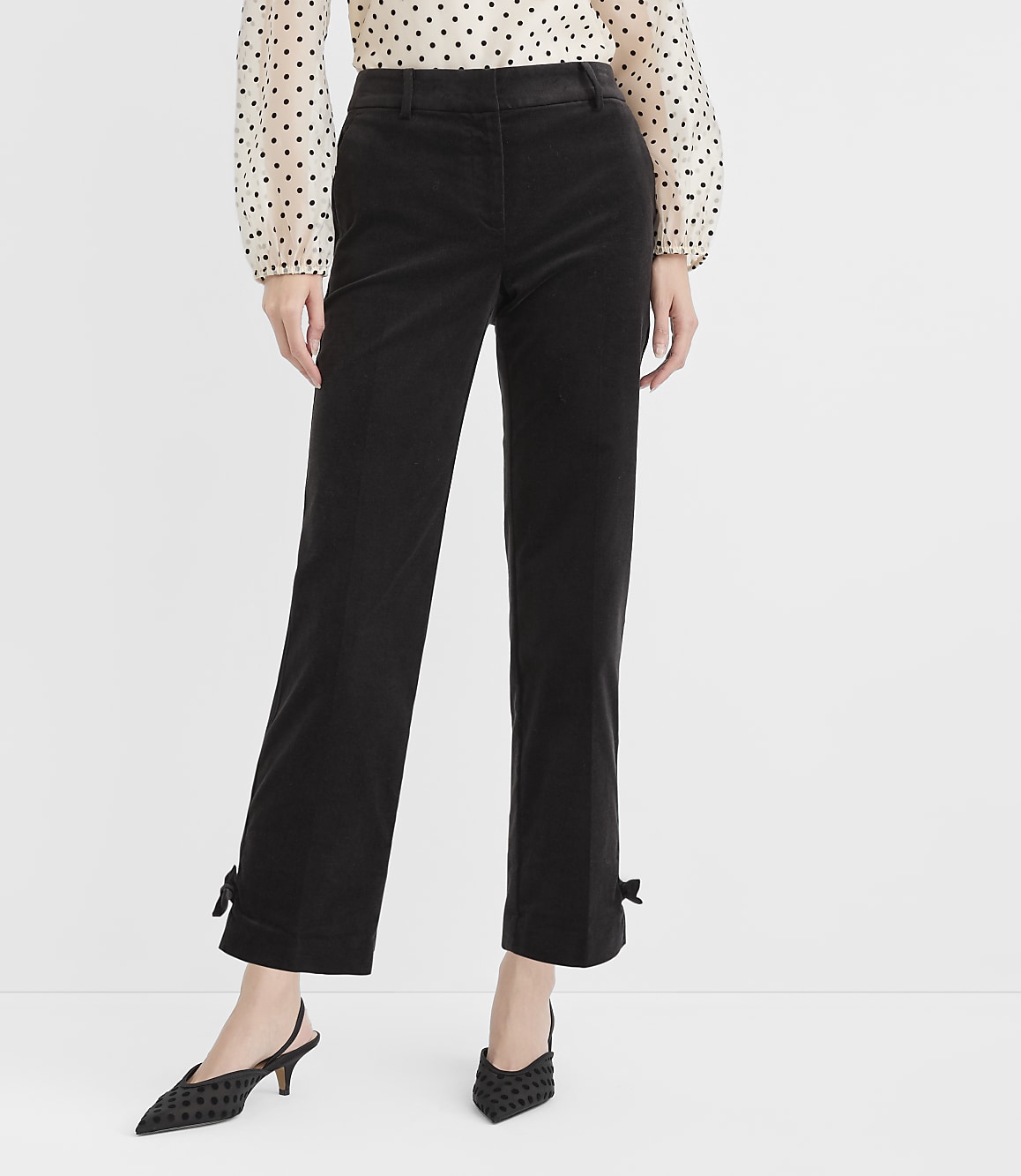 A person stands in front of a plain white background, wearing black corduroy pants with bows at the ankles and a polka-dot blouse.