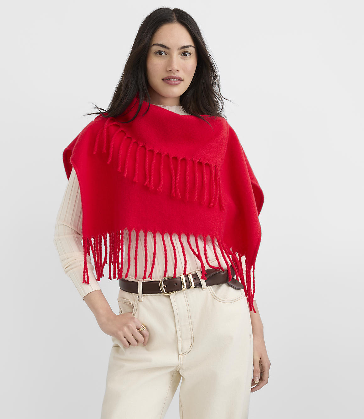 A woman in a cream sweater and light-colored pants wears a vibrant red fringed poncho against a plain white background.