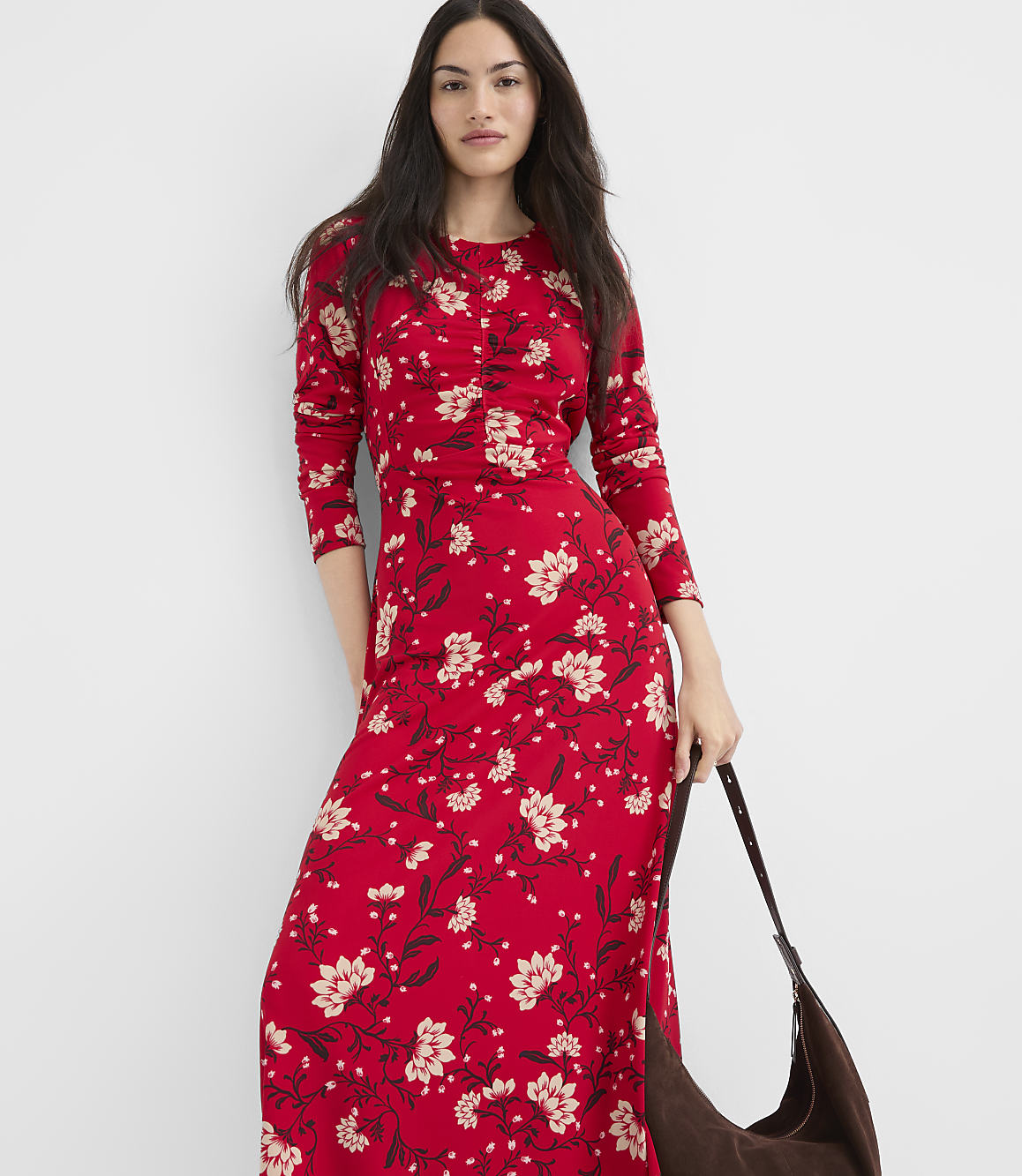 A woman in a red floral maxi dress stands against a plain white background, holding a brown shoulder bag.