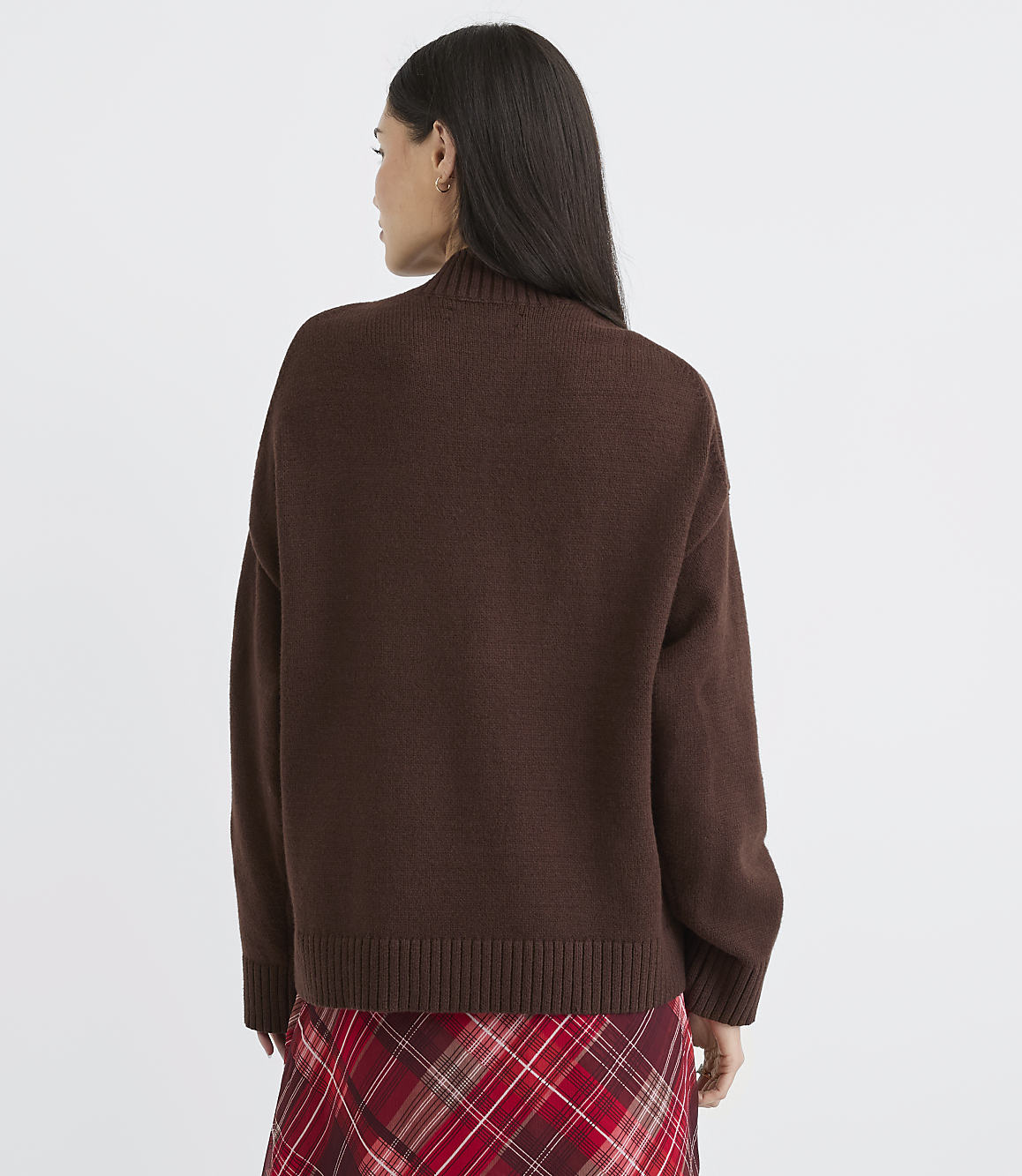 A woman in a brown sweater and plaid skirt stands against a plain white background.