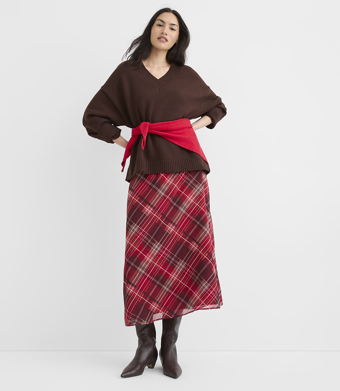 A woman in a brown v-neck sweater and a red plaid skirt stands against a plain white background.
