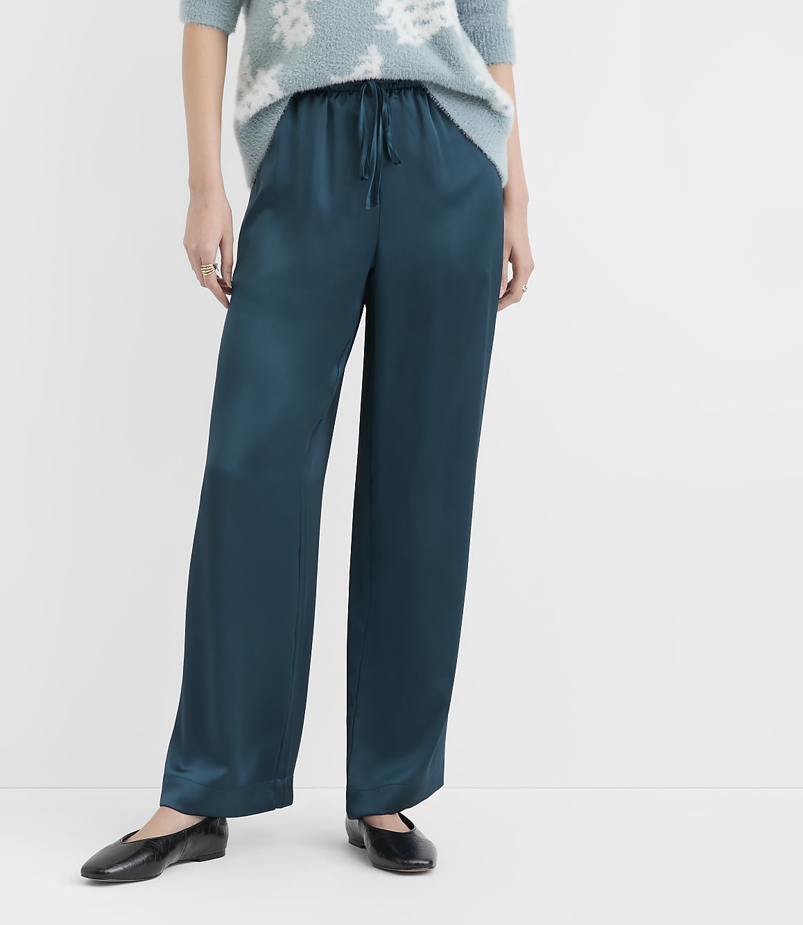 A person stands in teal satin pants and black flats against a plain white background.