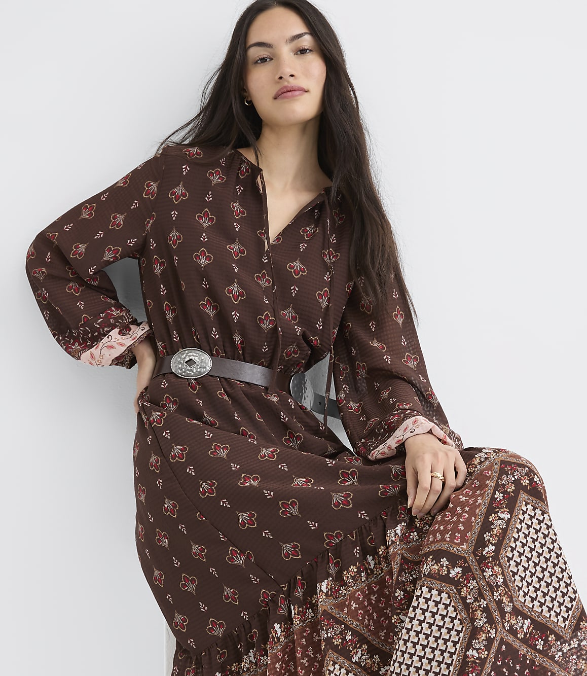 A woman in a patterned brown dress with a belt stands against a plain white background.