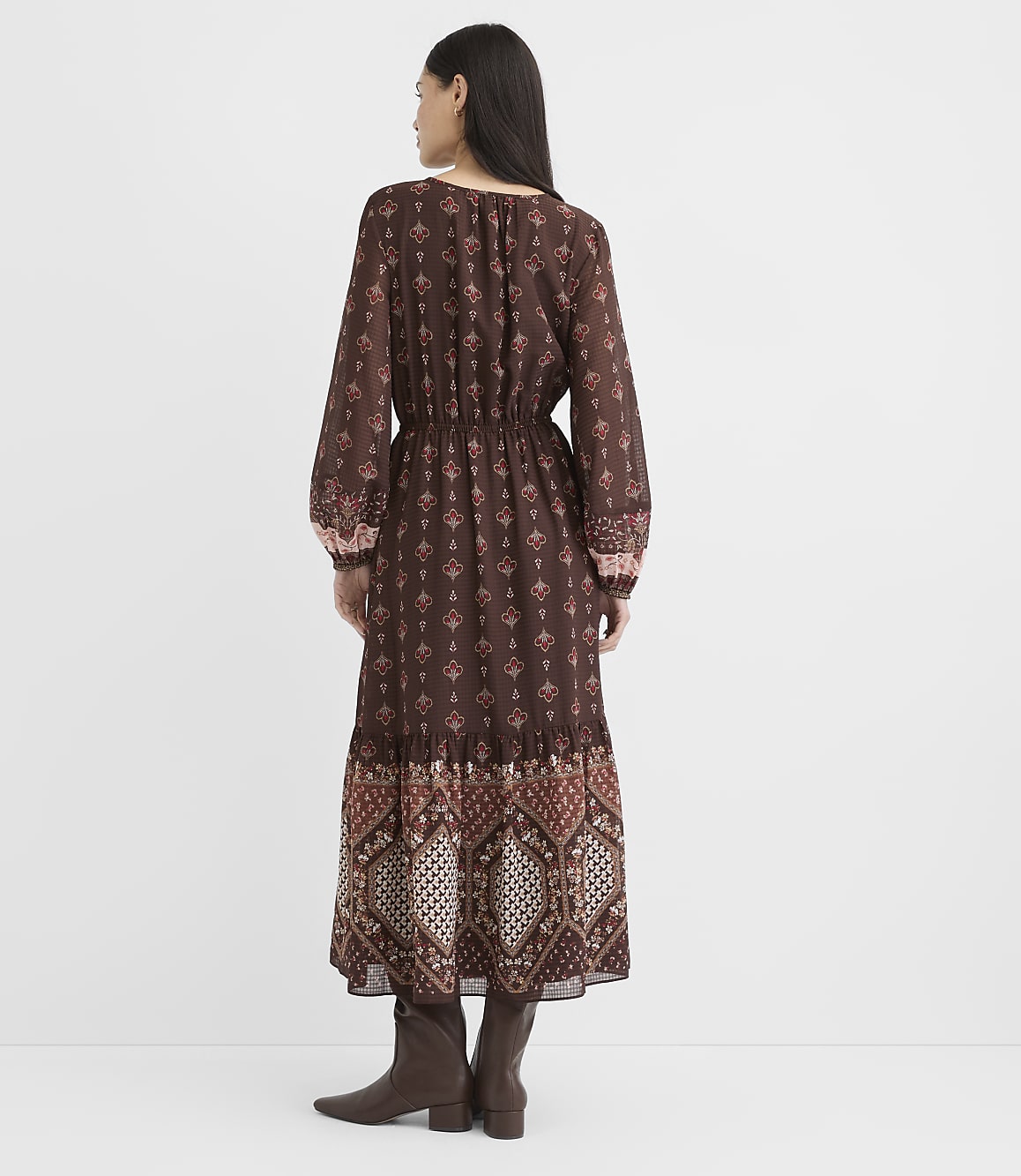 A woman in a long, patterned brown dress and boots stands against a plain white background.
