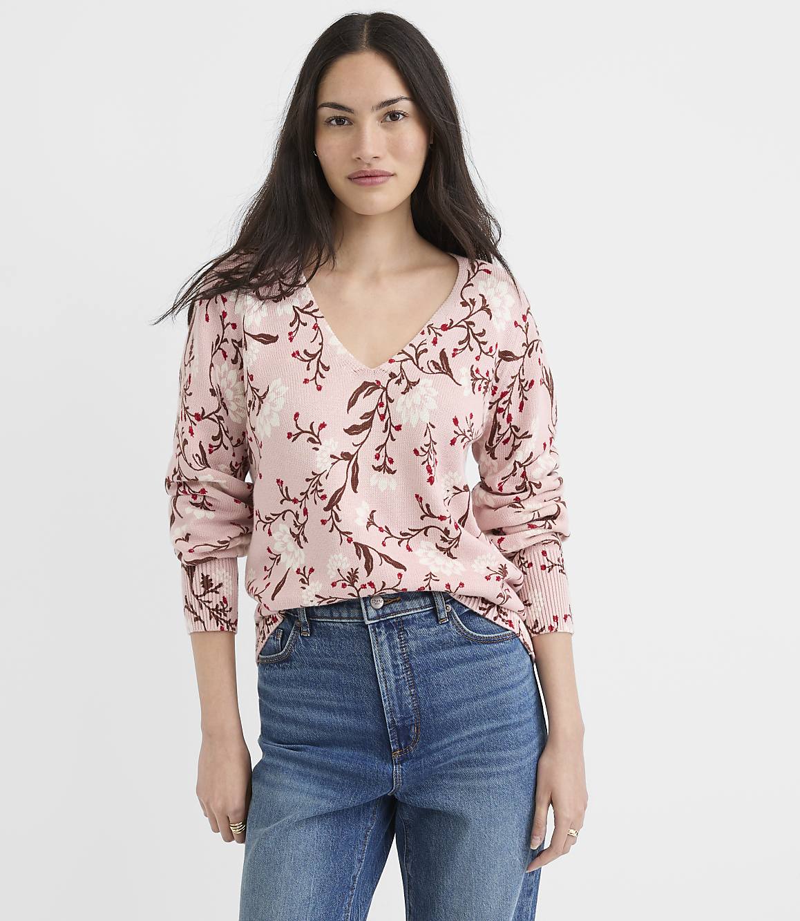 A woman in a floral pink sweater and blue jeans stands against a plain white background.