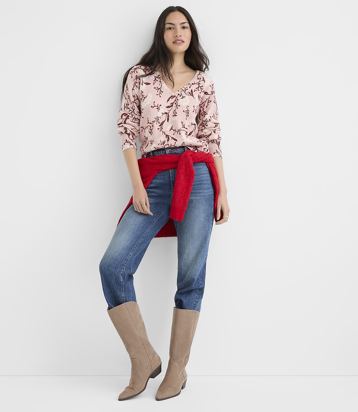 A woman in a floral sweater, jeans, and boots stands against a plain white background.