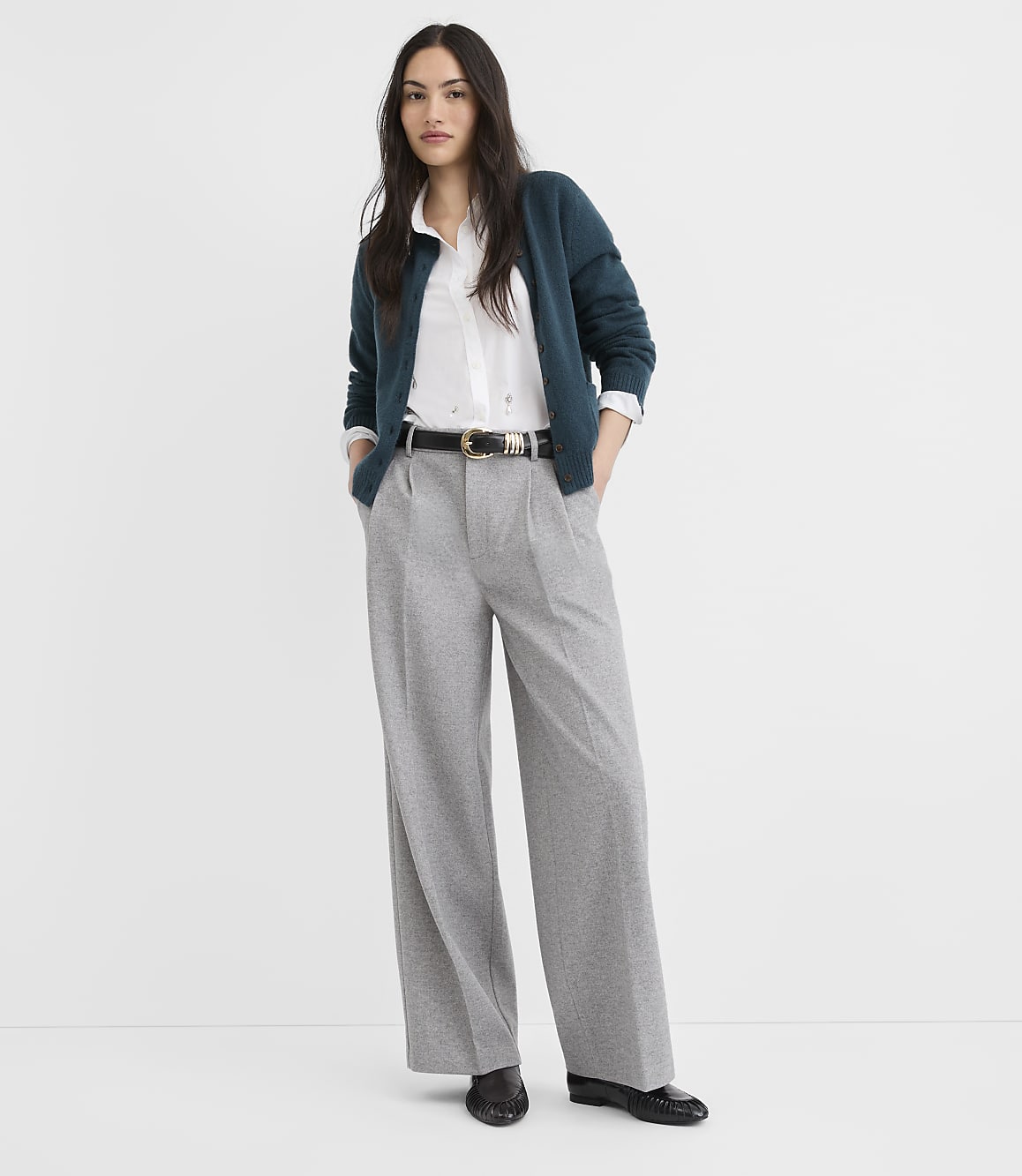 A woman in a teal cardigan and gray wide-leg pants stands against a plain white background.