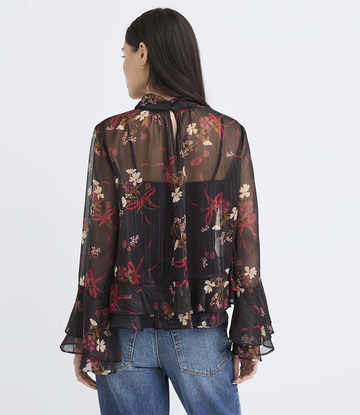 A woman in a sheer floral blouse and jeans stands against a plain white background.