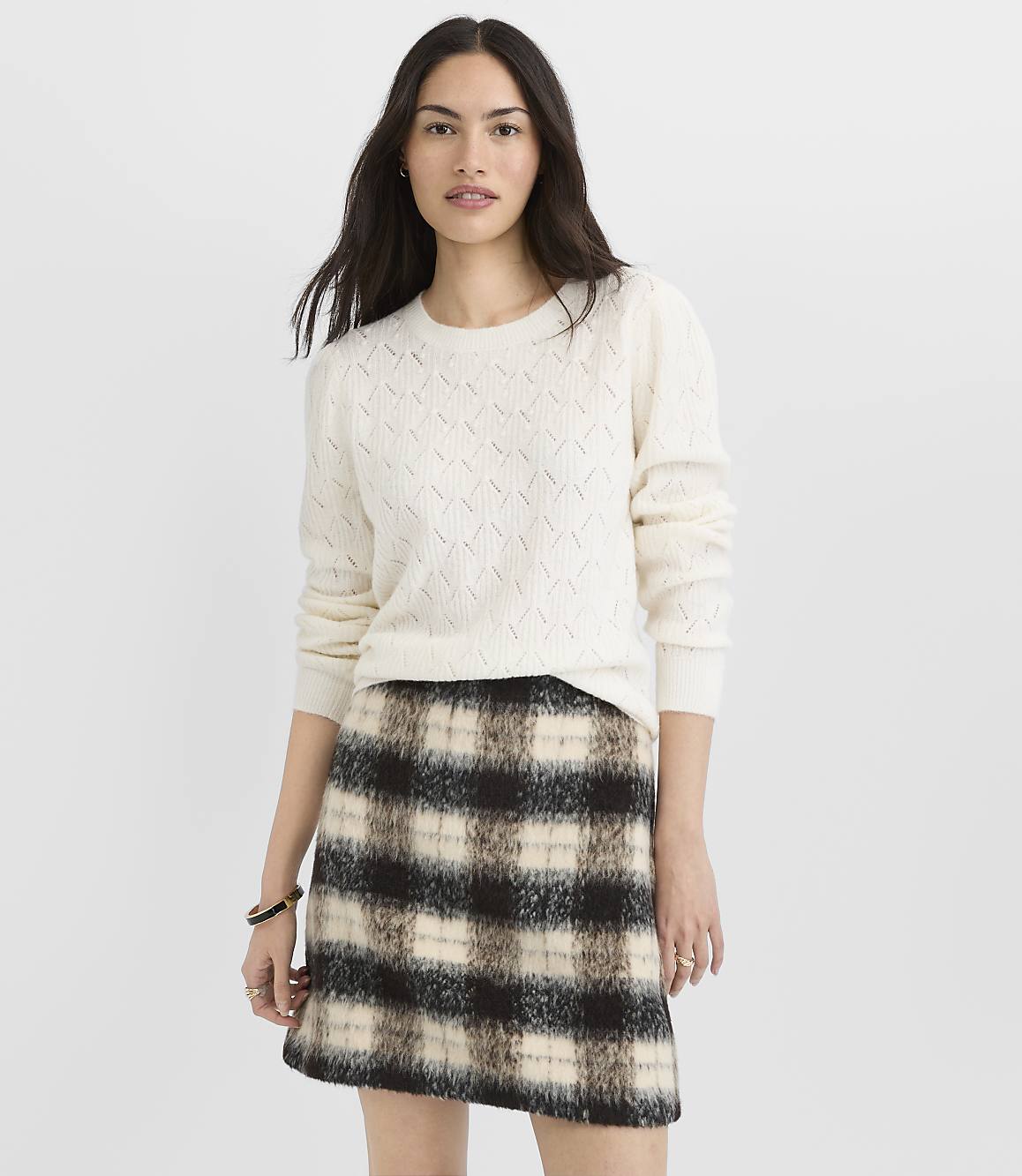 A woman in a cream cable-knit sweater and a plaid mini skirt stands against a plain white background.