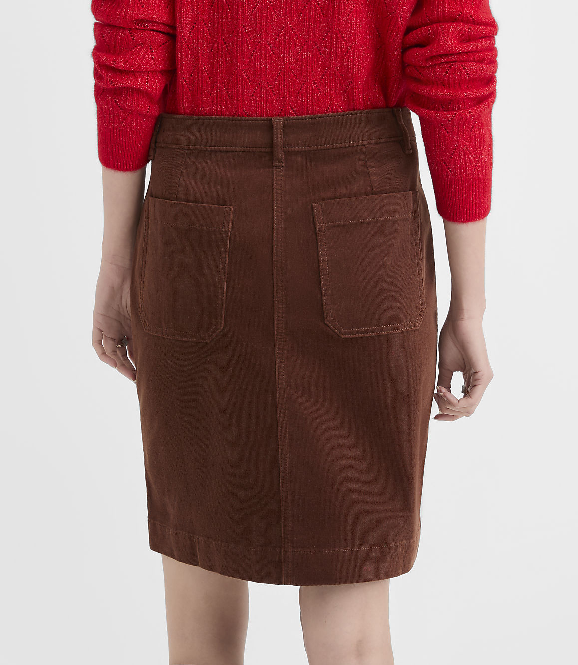 A person wearing a red sweater and a brown corduroy skirt stands against a plain white background.