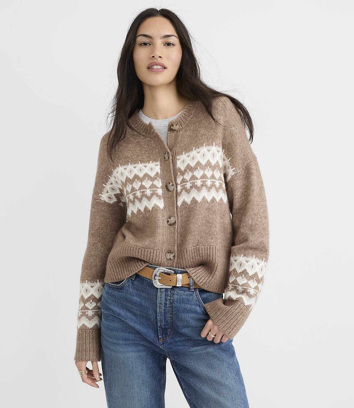 A woman in a brown and white patterned cardigan and blue jeans stands against a plain white background.