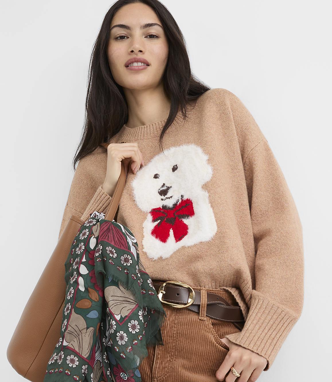 A woman in a tan sweater with a white dog applique and red bow, holding a brown tote bag with a patterned scarf, stands against a plain white background.