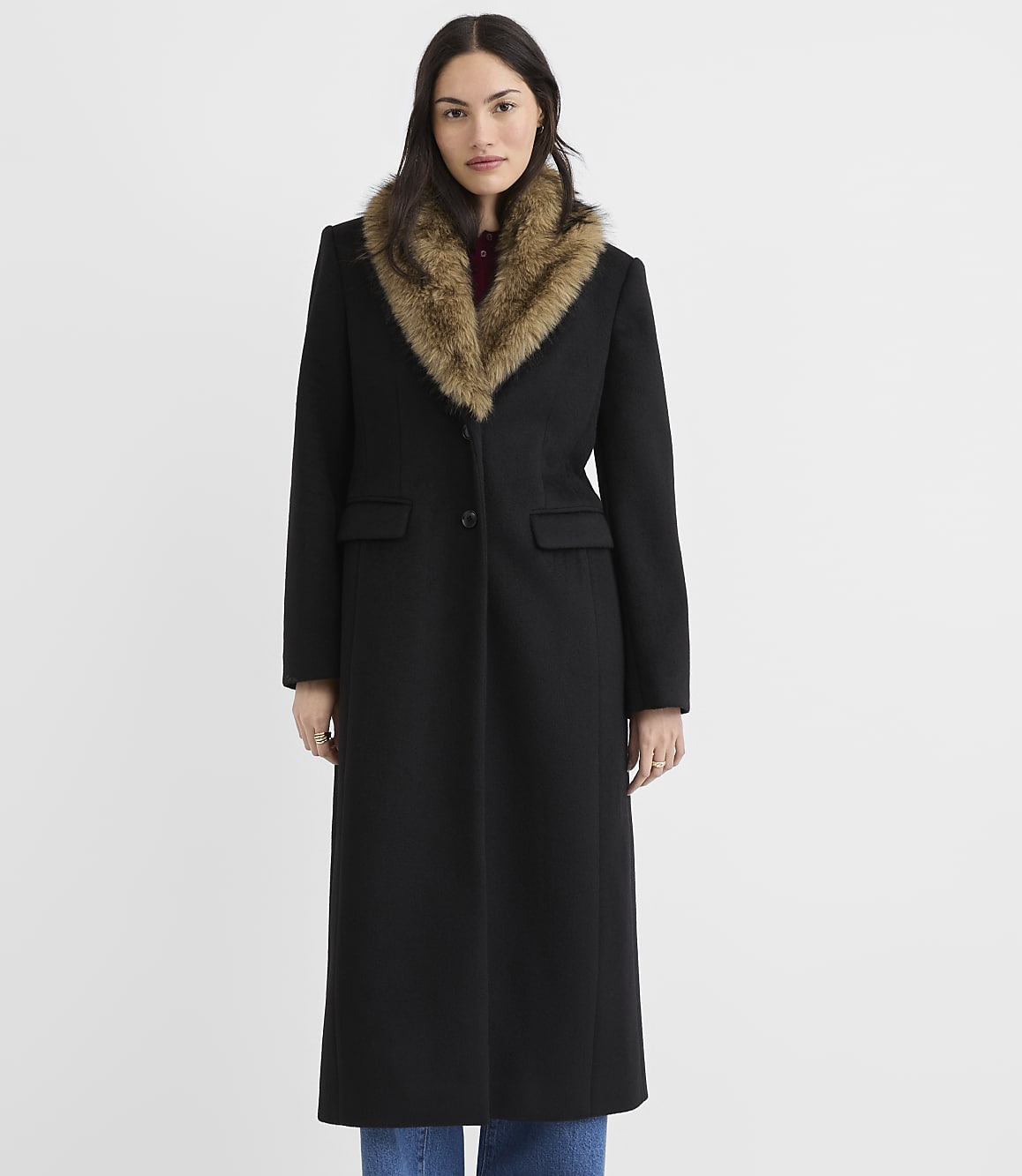 A woman in a long black coat with a fur collar stands against a plain white background.