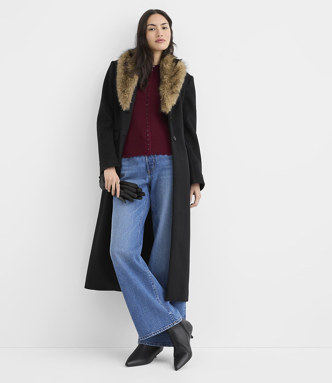 A woman in a long black coat with a fur collar, a burgundy top, and wide-leg jeans stands against a plain white background.