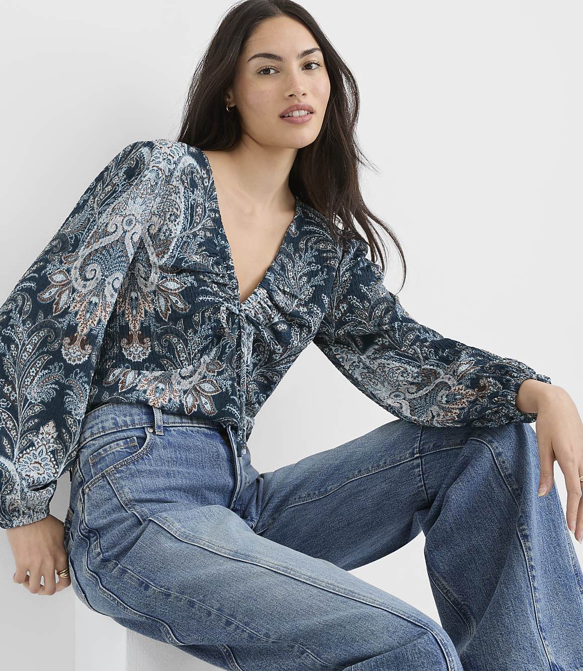 A woman in a patterned blouse and jeans sits against a plain white background.