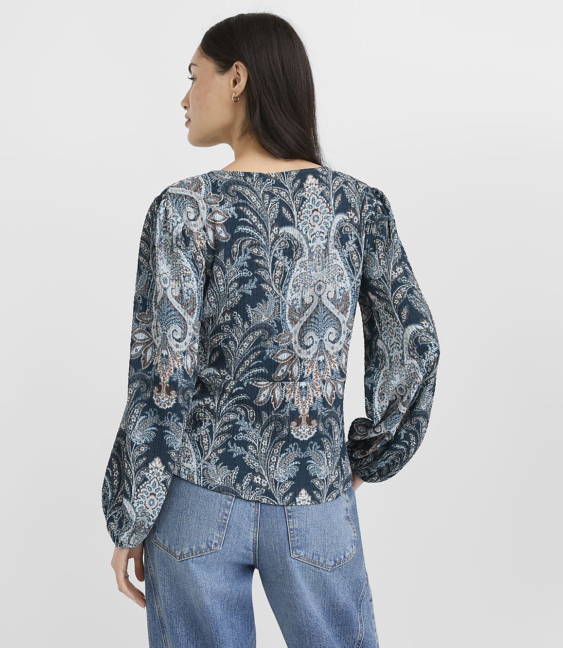 A woman in a patterned blouse and jeans stands against a plain background.