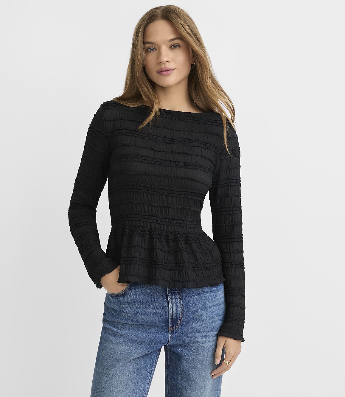 A woman in a black textured peplum top and blue jeans stands against a plain gray background.