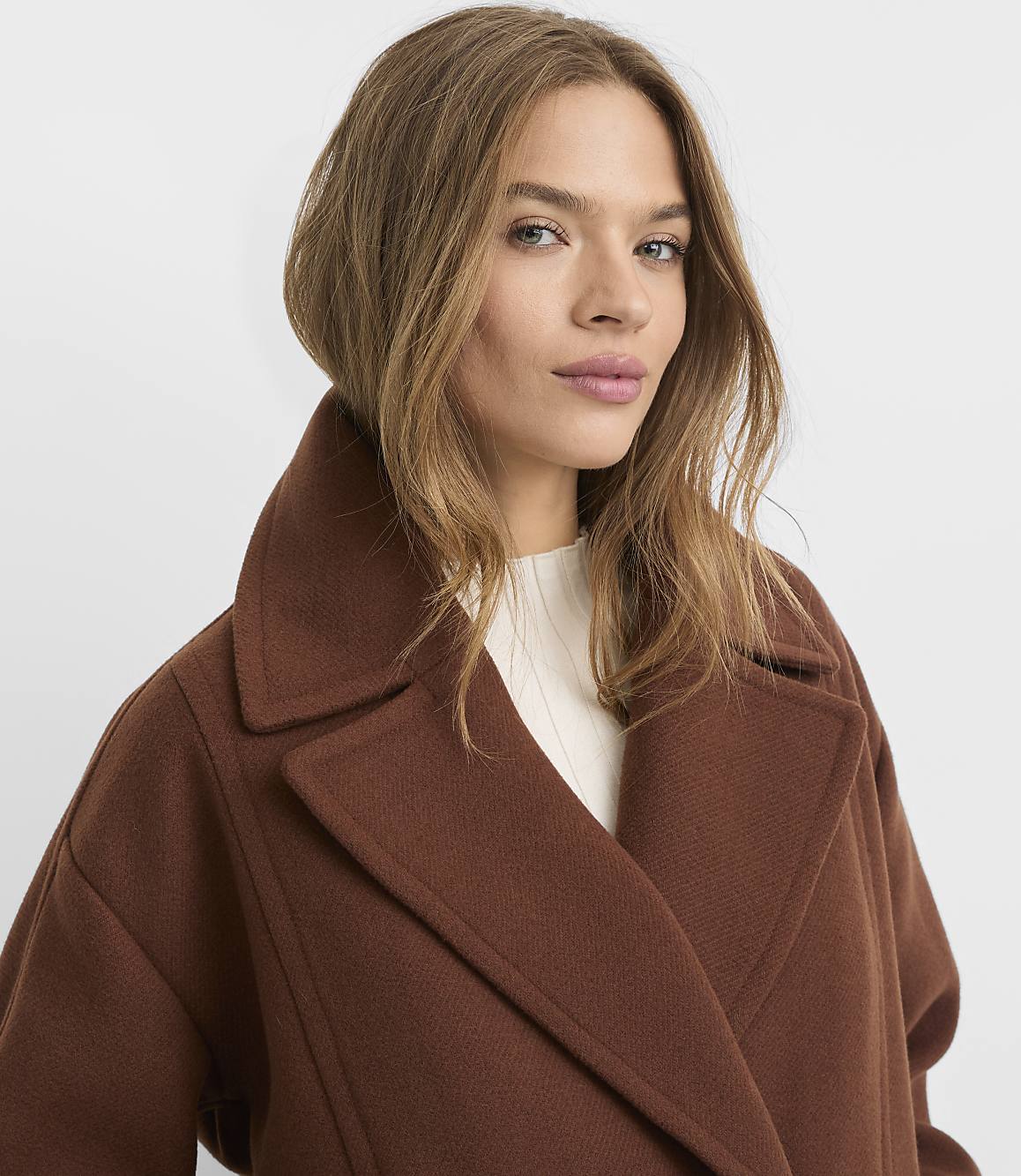 A woman in a brown coat with a white shirt is shown against a plain white background.
