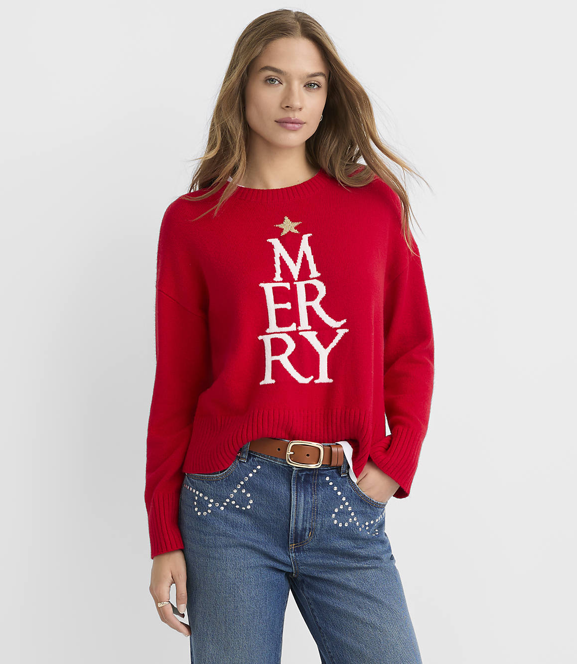 A woman in a red Christmas sweater and jeans stands against a plain white background.