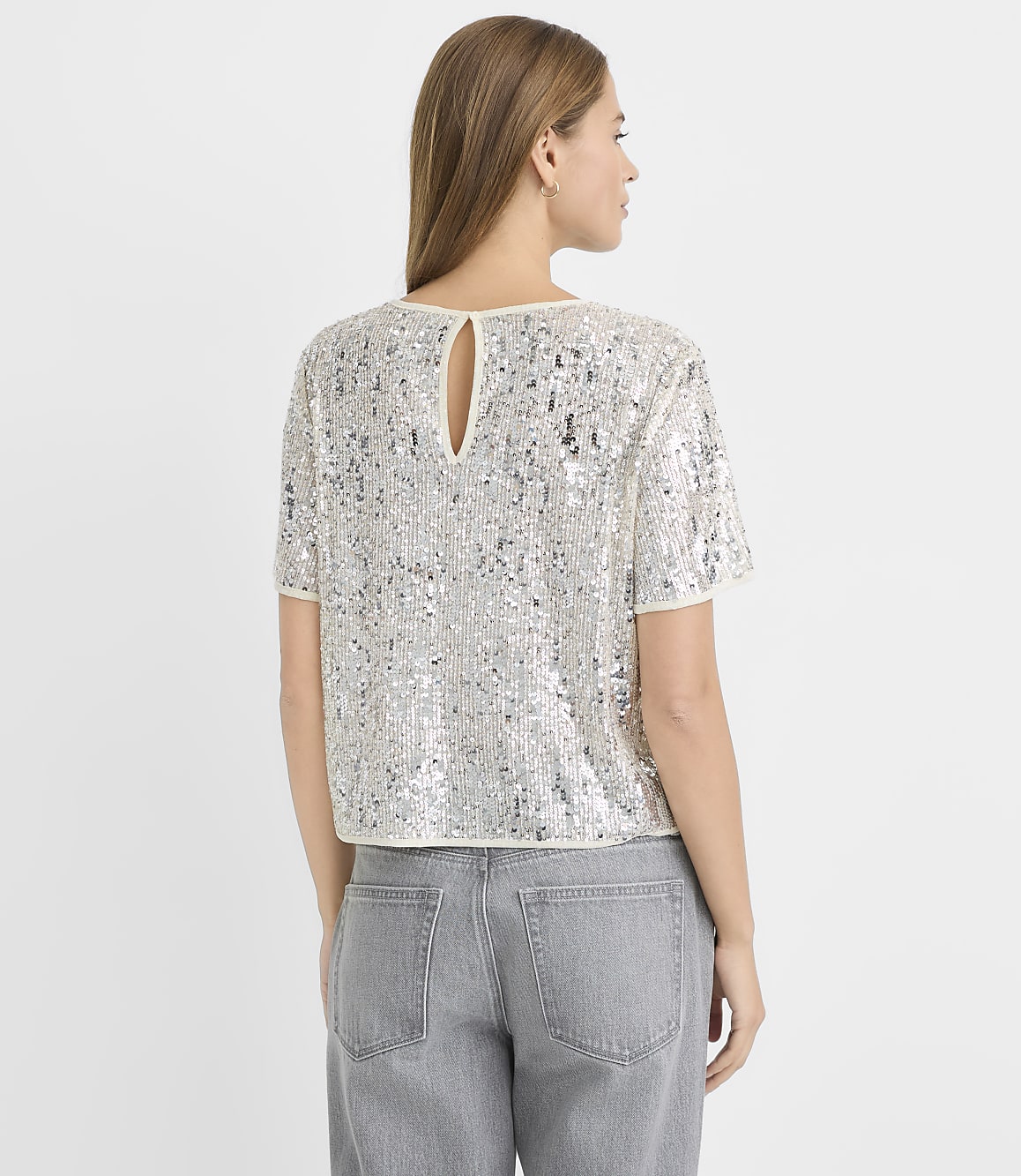 A woman in a sequined top and gray jeans stands against a plain white background.