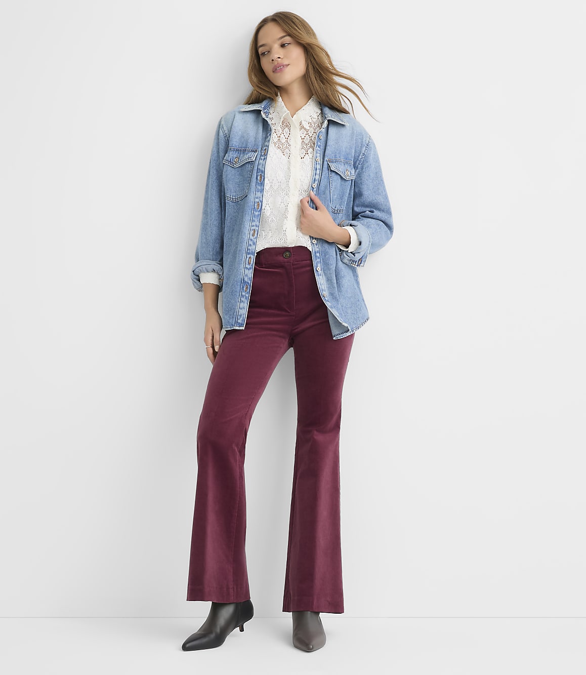 A woman in a denim jacket, white embroidered shirt, and burgundy flared pants stands against a plain white background.