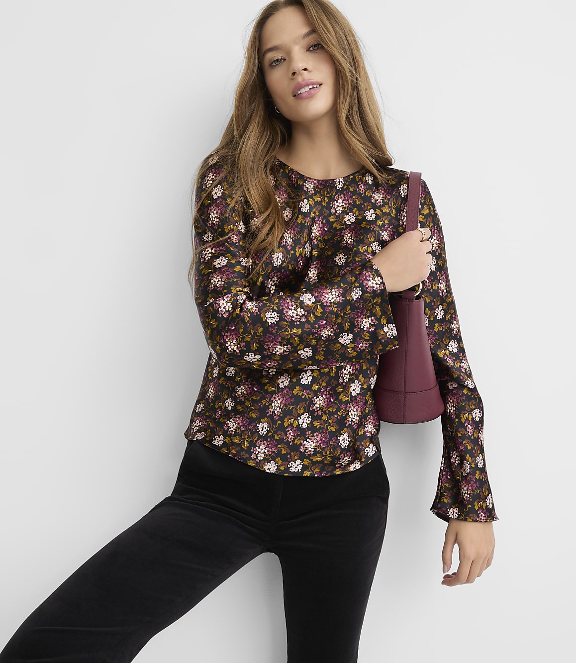 A woman in a floral top and black pants poses against a plain background, holding a burgundy purse.