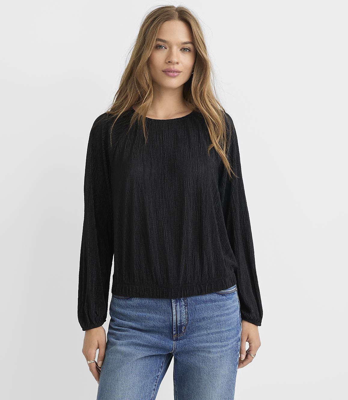 A woman in a black textured long-sleeved shirt and blue jeans stands against a plain white background.