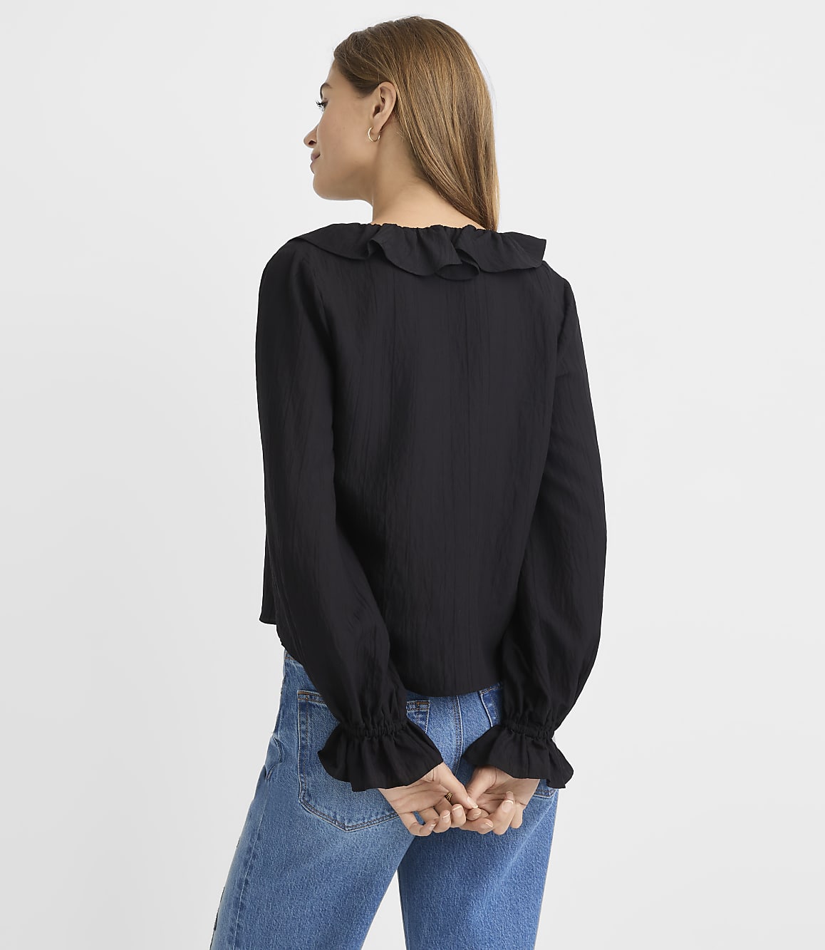 A woman in a black ruffled blouse and blue jeans stands against a plain white background.