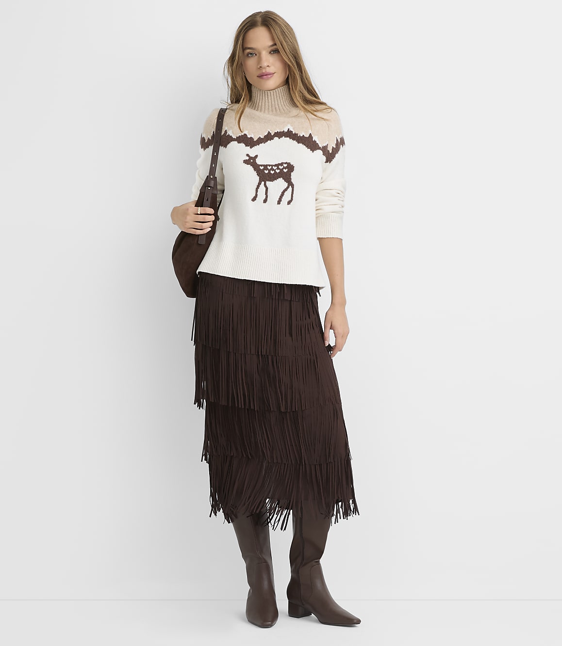 A woman in a cream sweater with a deer motif and a brown fringed skirt stands against a plain white background.