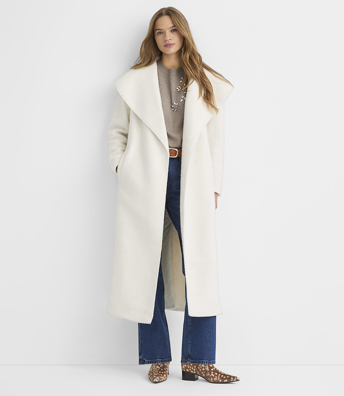 A woman in a long white coat and jeans stands against a plain white background.