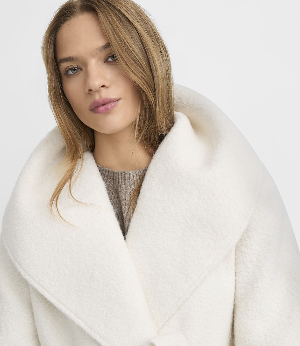 A woman with blonde hair and a neutral expression wears a cream-colored coat with a large collar, set against a plain white background.