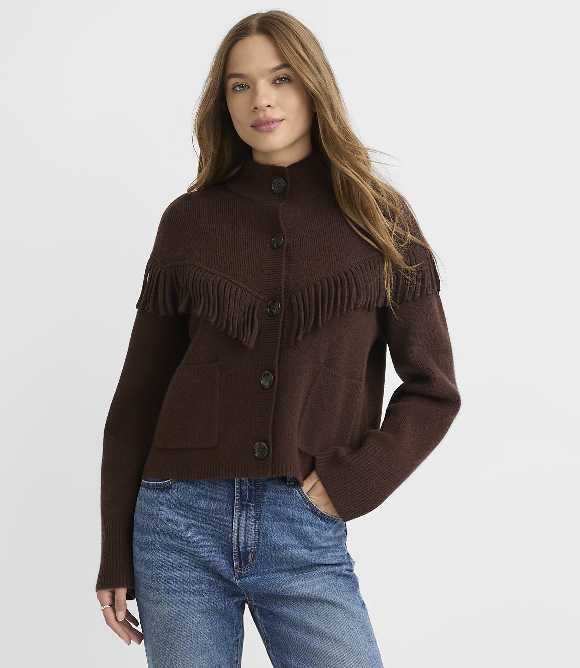 A woman in a brown fringed cardigan and blue jeans stands against a plain white background.