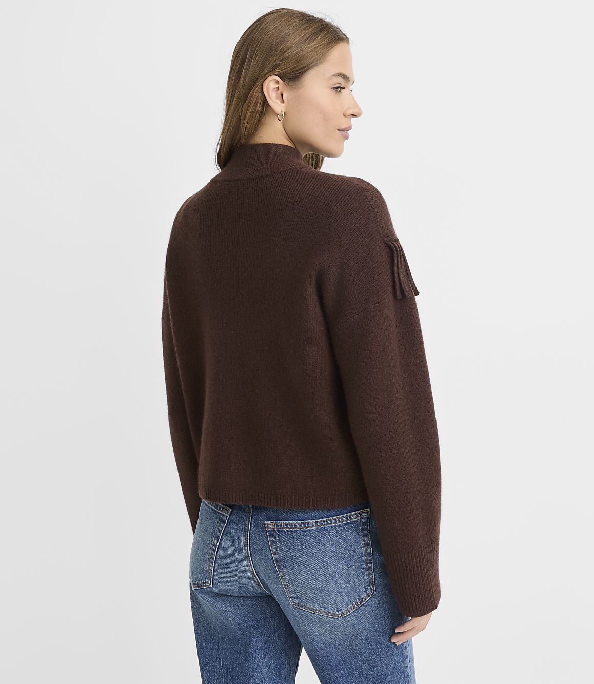 A woman in a brown sweater and blue jeans stands against a plain white background.