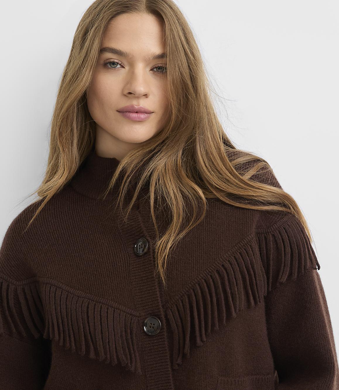 A woman with long, blonde hair wears a brown fringed cardigan against a plain white background.